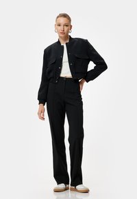 Zwarte cropped bomberjack met twee voorzakken, gecombineerd met zwarte rechte broek en witte sneakers, op een neutrale achtergrond.