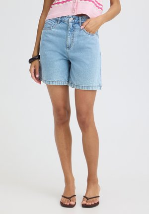 Frau trägt hellblaue Jeansshorts, rosa gestrickten Pullover mit wellenförmigem Muster, schwarzes Armband und braune Zehensandalen, steht mit der Hand in der Tasche.