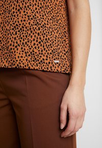 Hnědý top v barvě rzi s černými leopardími skvrnami, krátké rukávy a malá značka blízko lemu; spojený s hnědými strukturovanými kalhotami.