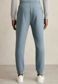 Pantaloni de trening albastre deschise, cu un talie elastică, picior conic și un buzunar pe spate, asortați cu adidași albi pe un fundal neutru.