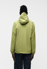 Άνδρας με κοτσίδες ντυμένος με ανοιχτοπράσινο μπουφάν με κουκούλα και σκούρα τζιν με κόκκινη κεντητική, στέκεται γυρισμένος με την πλάτη σε λευκό φόντο.