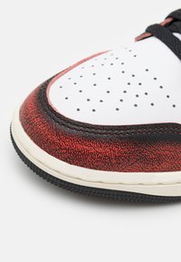 Gros plan sur l’avant d’une basket avec cuir blanc perforé, superposition texturée rouge, bordure noire, lacets noirs et semelle blanche.