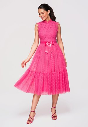 Kvinde i en ærmeløs, lys pink blonde- og tyl-middellang kjole med satinbånd i taljen, poserer med den ene arm strakt ud og kigger ned.