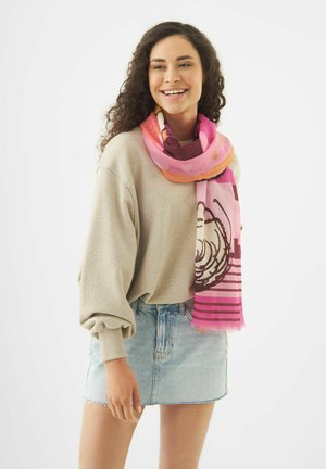 Jonge vrouw glimlachend, draagt een beige oversized trui, lichtblauwe spijkerrok en een roze sjaal met patroon om haar hals.