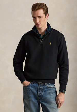 Jeune homme portant un pull noir à fermeture éclair quart de zip avec un logo jaune, une chemise à carreaux bleue en dessous, un jean bleu et une montre-bracelet.