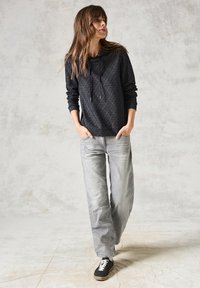 Sort mønstret hoodie med snore, løstsiddende grå jeans og sorte sneakers. Baggrunden har en tekstureret, lysfarvet væg.