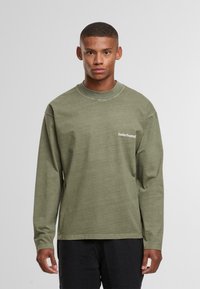 Jauns vīrietis ar īsiem matiem, ģērbts olīvzaļā garajā piedurknē T-kreklā ar tekstu "SundayRunning" un melnām biksēm, stāv uz neitrāla fona.