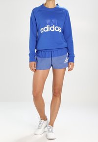 Sudadera azul con el logo de "adidas", combinada con shorts a rayas azules. Presenta un material ligero y un diseño atlético. Se lleva con zapatillas blancas.
