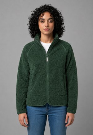 Kvinde med krøllet hår iført en grøn tekstureret fleecejakke med lynlås og blå jeans, stående mod en grå baggrund.