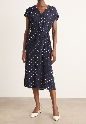 Femme portant une robe midi bleu marine à pois blancs, manches courtes et taille nouée, associée à des talons blancs à bouts pointus.