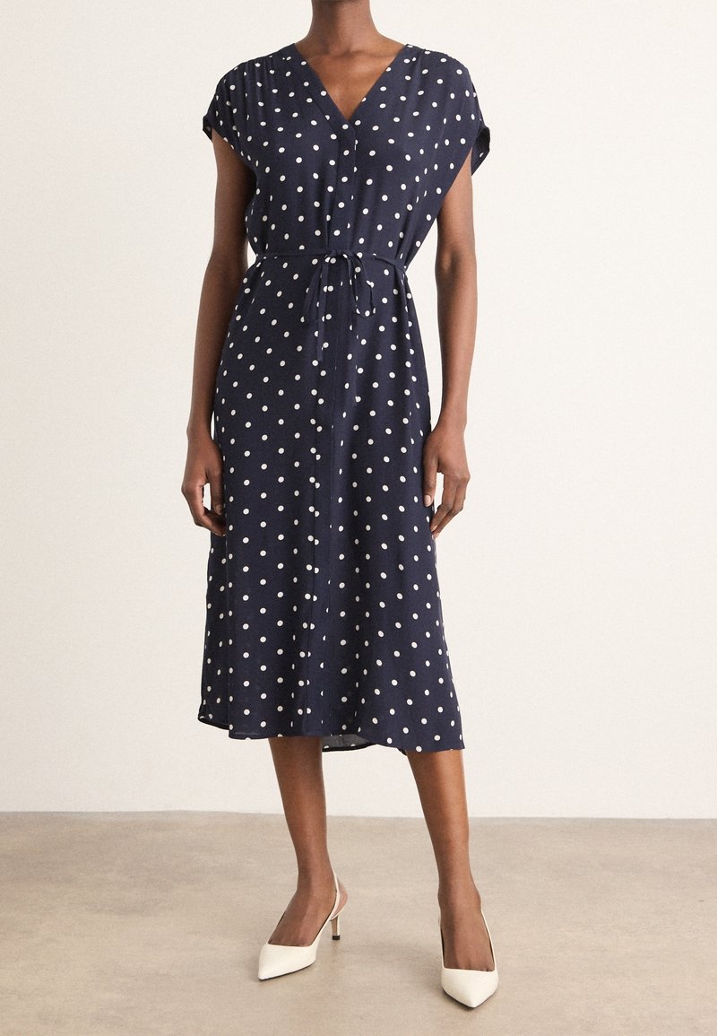 Femme portant une robe midi bleu marine à pois blancs, manches courtes et taille nouée, associée à des talons blancs à bouts pointus.