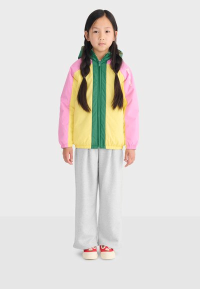Jeune fille aux longues tresses portant une veste color-block en jaune, rose, vert, un pantalon gris et des baskets rouges ornées de motifs floraux.