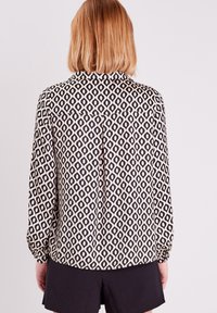 Blouse à motif noir et crème avec des manches longues, arborant un design en diamant et un détail de dos boutonnière. Tissu lisse, coupe décontractée.