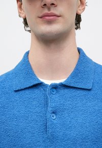 Jovem a usar uma camisa azul texturada com colarinho abotoado, com uma t-shirt branca visível na gola, contra um fundo simples.