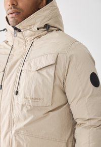 Giacca con cappuccio beige, dotata di tasche sulle spalle e sul petto, tessuto testurizzato, cordoni regolabili e una toppa circolare nera sulla manica.