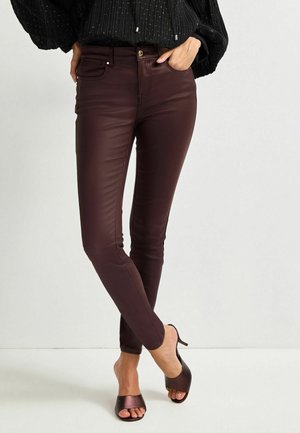 Femme portant un pantalon en cuir marron foncé slim et des chaussures à talons hauts ouvertes marron, debout avec une jambe croisée devant l'autre.