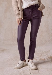 Lila figurnära byxor gjorda av ett glänsande, slätt material. Har en traditionell femfickdesign och knappstängning fram. Bärs med vita sneakers.
