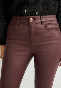 Prim-plan al unei persoane care poartă pantaloni din piele bordeaux cu talie înaltă, cu detalii din butoane și nituri aurii, asociați cu un top texturat negru.