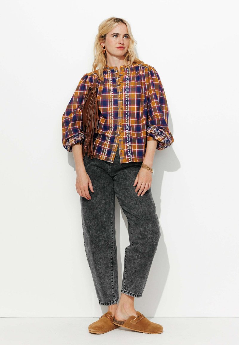 Chemise à carreaux orange, violet et jaune avec des manches bouffantes et une finition en dentelle, associée à un jean gris foncé et des sabots marron. Les accessoires comprennent un sac à franges.