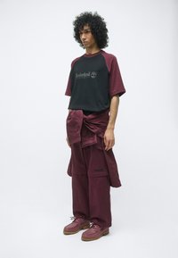 Zwarte en bordeauxrode Timberland t-shirt, bordeauxrode cargobroek en bijpassende schoenen. De stof lijkt glad; het shirt heeft een logo; gelaagd ontwerp zichtbaar.