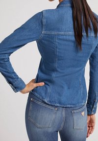 Giubbotto in denim blu con vestibilità slim, maniche lunghe e polsini abbottonati. Dettagli a toppa sulla schiena e sulle tasche. Indossato con jeans in denim abbinati.