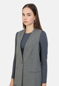 Grå väst med skräddarsydd design, med en knapp framtill, två fickor och en slät textur över en mörk långärmad topp.