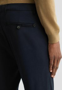 Marineblaue Hose aus glattem Stoff, mit einem Reißverschluss-Rückentasche und Knopfverschluss sowie einer taillierten Passform. Sichtbare Hand an der Seite.