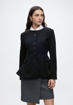 Femme portant un blazer noir boutonné avec un détail péplum sur une chemise blanche et une jupe asymétrique grise, debout devant un fond uni.