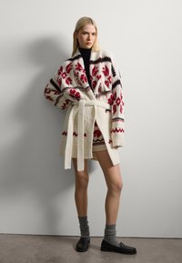 Cárdigan de punto con patrones florales rojos sobre un fondo marfil, cintura ceñida con cinturón, escote en V profundo y mangas oversized. Combinado con calcetines grises y mocasines negros.