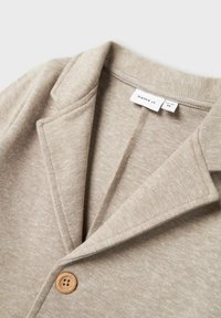 Felpa beige con colletto a taglio, tessuto testurizzato e chiusura con un singolo bottone in legno. L'etichetta recita "name it" sul collo.
