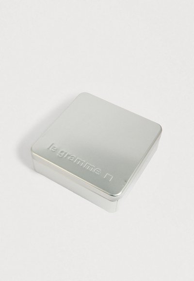 Kompakta sudraba metāla kaste ar noapaļotām malām un izcelto "le gramme" tekstu uz vāka, novietota uz baltas virsmas.