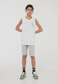 Camicetta senza maniche in cotone bianca con tasca grafica "LA", abbinata a pantaloni grigi. Presenta scarpe da ginnastica in bianco e nero con calzini a righe.