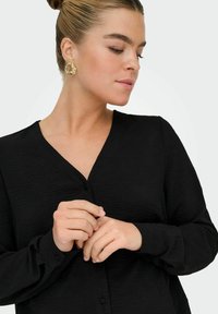 Blouse noire à manches longues, avec un décolleté en V et un tissu texturé. Des boucles d'oreilles en or en forme de créole sont visibles en accent.