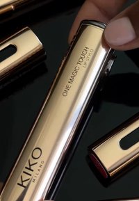 KIKO Milano One Magic Touch Lip Stylo, joka on kiiltävässä kultametallisessa putkessa, jossa on kaiverrettu brändäys ja tyylikäs muotoilu, lepäävä heijastavalla pinnalla.
