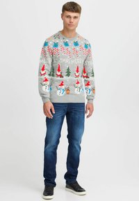 Grauer Weihnachtspulli mit leuchtenden Farben, Schneemännern, Bäumen und Geschenken in einem verspielten Design. Kombiniert mit dunklen blauen Jeans und schwarzen Schuhen.