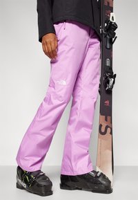 Roze waterdichte skibroek met een losse pasvorm, zijzakken en een klein wit logo. Zwarte skischoenen met groene accenten maken de outfit compleet.