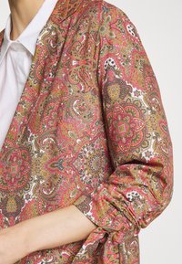 Blouse colorée à motifs avec des dessins cachemire rouges, roses et dorés. Présente une coupe décontractée et des manches froncées, fabriquée en tissu léger.