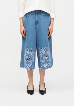 Žena nosi kratke široke plave traperice s bijelim cvjetnim vezom blizu valovitog ruba, uparene s crnim šiljastim cipelama.