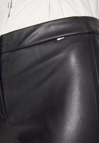 Gros plan d'une personne portant une chemise blanche rentrée dans un pantalon en cuir noir taille haute avec une petite étiquette en tissu tricolore sur la ceinture.