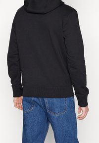 Felpa nera con cappuccio a coulisse, tasca a marsupio, polsini e orlo a costine, abbinata a jeans di denim blu. Tessuto liscio, vestibilità casual.