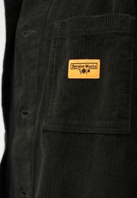 Cămașă din velur verde închis cu o etichetă galbenă "Service Works" pe buzunarul de la piept. Prezintă o suprafață texturată și închideri cu nasturi.
