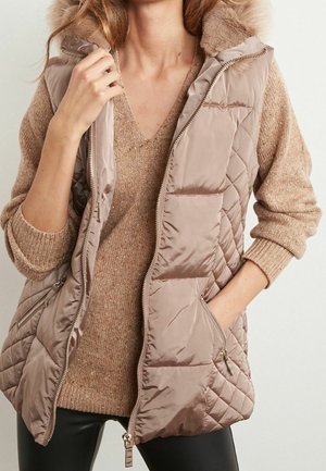 Femme portant une veste matelassée beige avec col en fourrure sur un pull beige en maille à col en V et un pantalon noir, main dans la poche de la veste, fermeture éclair partiellement ouverte.