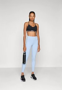 Fekete sportmelltartó fehér logóval, világoskék magas derekú leggings, fekete sportcipő. Fekete vízálló palackot tart a kezében.