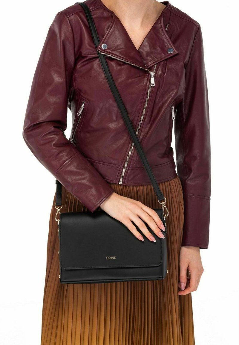 Czarna torebka na ramię o eleganckim, prostokątnym kształcie, gładkiej fakturze i złotymi detalami logo, trzymana w dłoni w zestawieniu z plisowaną spódnicą.