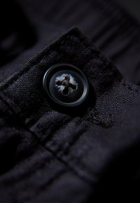 Zbliżenie czarnego guzika z czterema otworami, przyszytego ciemną nicią na fakturowanej czarnej tkaninie, podkreślające szycie i splot tkaniny.