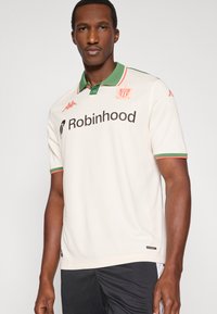 Kreminės spalvos polo marškinėliai su žalia apykakle ir raudonomis detalėmis, ant kurių yra užrašas „Robinhood“ ir logotipas. Pagaminti iš tekstūruotos medžiagos, trumpomis rankovėmis.