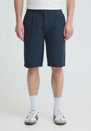 Mand iført marineblå skræddersyede shorts, hvide strømper og hvide sneakers med sorte striber, stående mod en neutral baggrund.