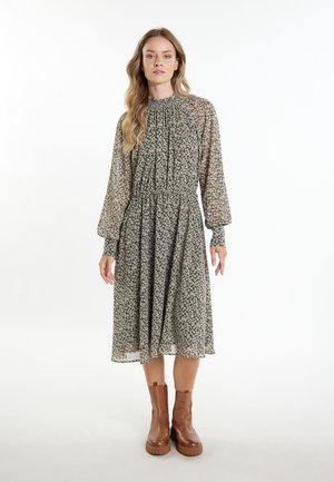 Vrouw met lang golvend haar, een lange mouwen bloemen midi-jurk en bruine enkellaarsjes dragend, staand voor een witte achtergrond.