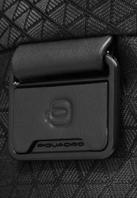 Materiale nero testurizzato con una fibbia rettangolare nera con il logo Piquadro, su uno sfondo decorato.