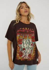 Brązowy t-shirt graficzny z vintage'owym nadrukiem przedstawiającym trzy białe konie oraz kwiatowe akcenty w kolorze czerwonym i zielonym. Luźny krój z krótkimi rękawami.